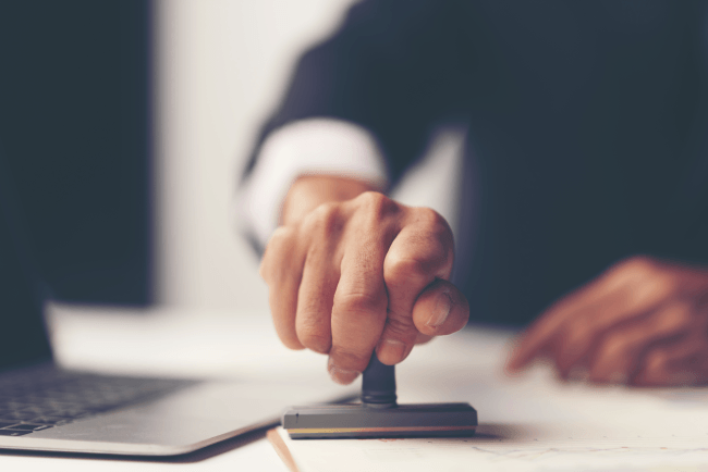 Close-up of a person's hand stamping with approved stamp on document at desk